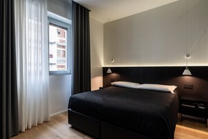 A modern bedroom with a large bed, a nightstand, a wall-mounted lamp, and a window with curtains.