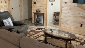 A cozy living room with a leather sofa, a glass coffee table, and a wooden floor. There is a television mounted on the wall and a door leading to another room. - Cozy cabin in the woods.   Close to boat ramp.  Shared 5 bay boat storage  (Counce)