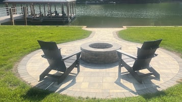 Un espace extérieur circulaire aménagé avec des sièges, un foyer, deux chaises de jardin et un belvédère situé au bord d’un lac.