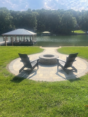 Un espace extérieur circulaire aménagé avec des sièges, un foyer, deux chaises de jardin et un belvédère situé au bord d’un lac.