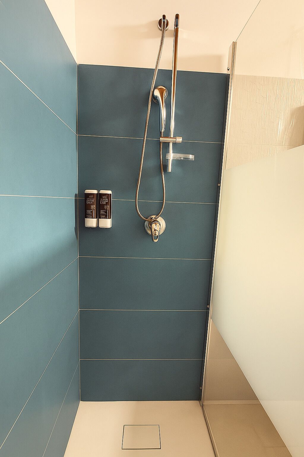 A modern bathroom with a blue tiled wall, a shower with a handheld showerhead, and two bottles of shampoo and conditioner on the wall.