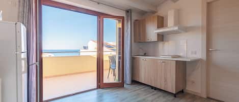 A modern kitchen with wooden cabinets, a white countertop, and a sliding glass door leading to a balcony with a view of the sea.