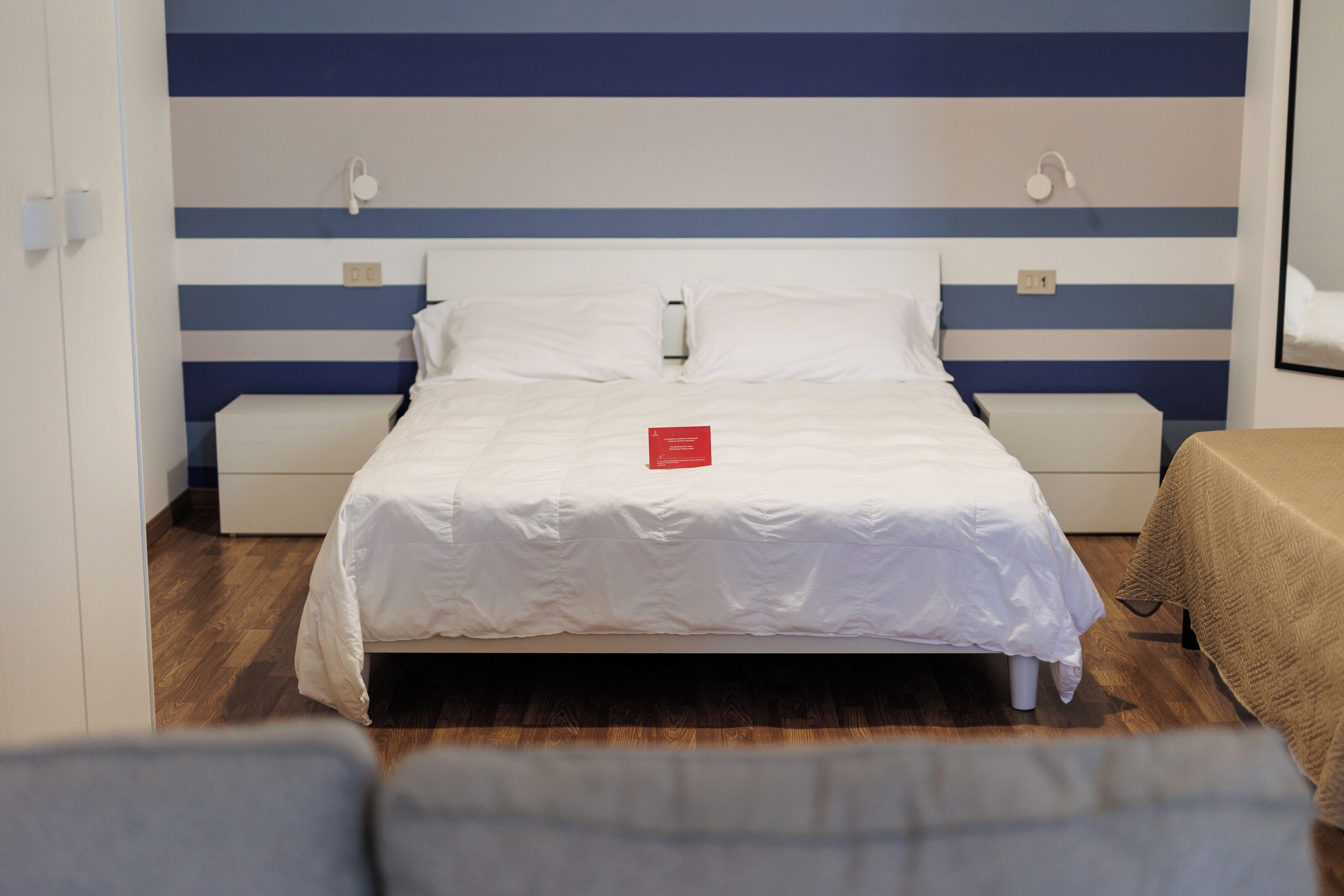 Una cama bien hecha con ropa blanca, un libro rojo encima y una pared a rayas al fondo.