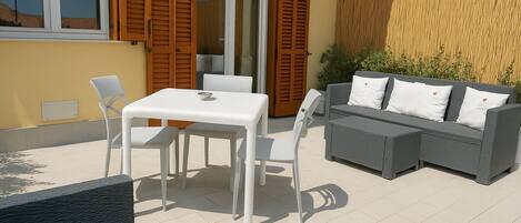 An outdoor patio with a white table and chairs, a grey sofa with white cushions, and a wooden fence.