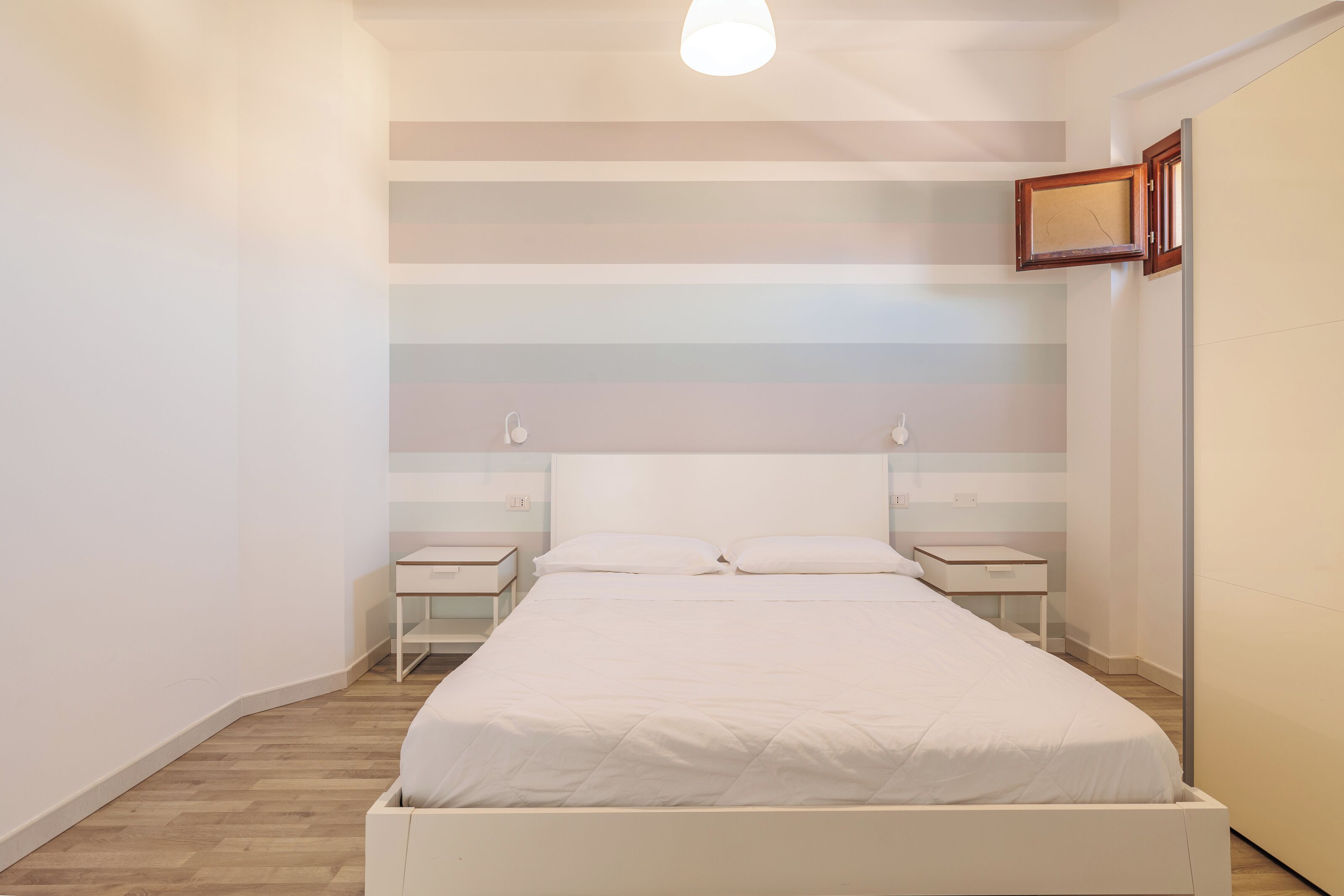 A bedroom with a bed, two bedside tables, a nightstand, a wooden floor, and a striped wall.