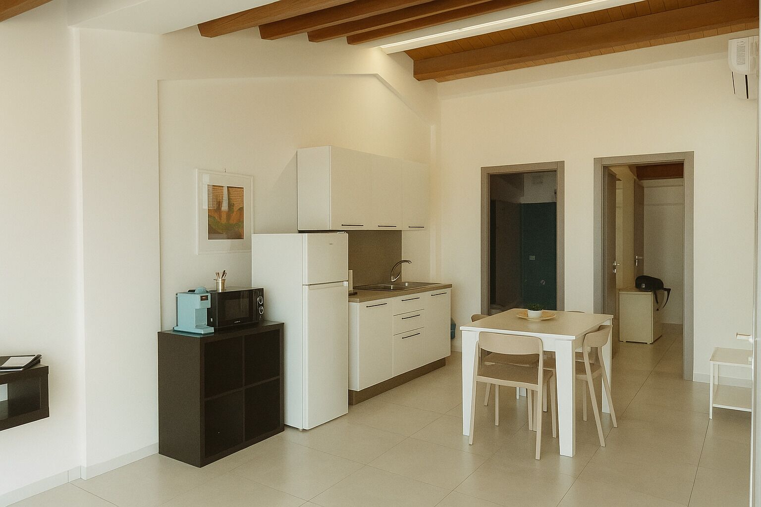 Cocina compacta con armarios blancos, microondas y refrigerador. Hay una mesa de comedor con sillas y un mueble pequeño.