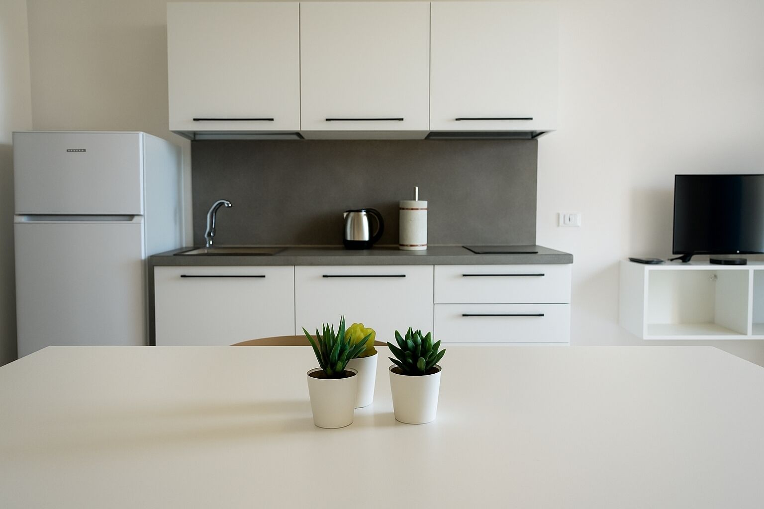 A modern kitchen with white cabinets, a grey countertop, and a white dining table. There are two potted plants on the table.