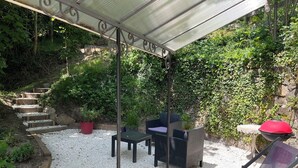 A covered patio with outdoor furniture, a grill, and a staircase leading to a garden area.