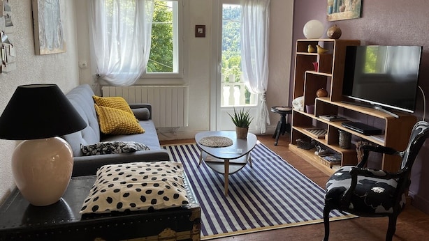 A living room with a sofa, a coffee table, a TV stand, and a chair. There are curtains on the windows, and a plant on the coffee table.