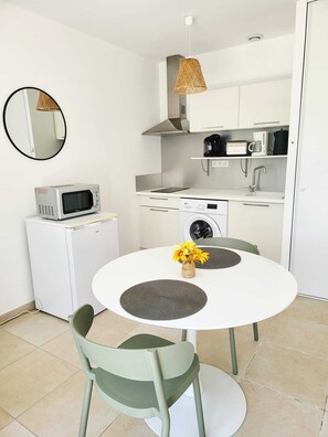 Uma cozinha compacta com micro-ondas, torradeira e máquina de lavar, uma mesa redonda com duas cadeiras e um lustre.