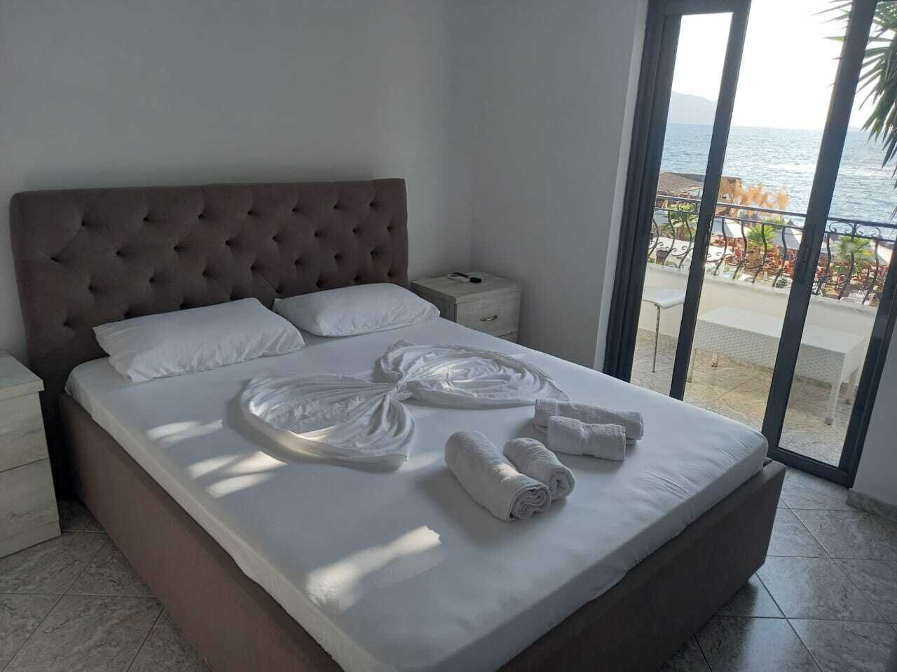 A neatly made bed with towels arranged in a fan shape, a tufted headboard, and a view of the sea through a glass door.