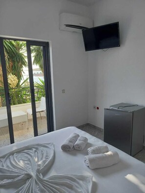 Ein Hotelzimmer mit Bett, flachbildfernseher an der Wand, kleinem Kühlschrank und einer Glastür, die zu einem Außenbereich mit einer Palme und Meerblick führt.