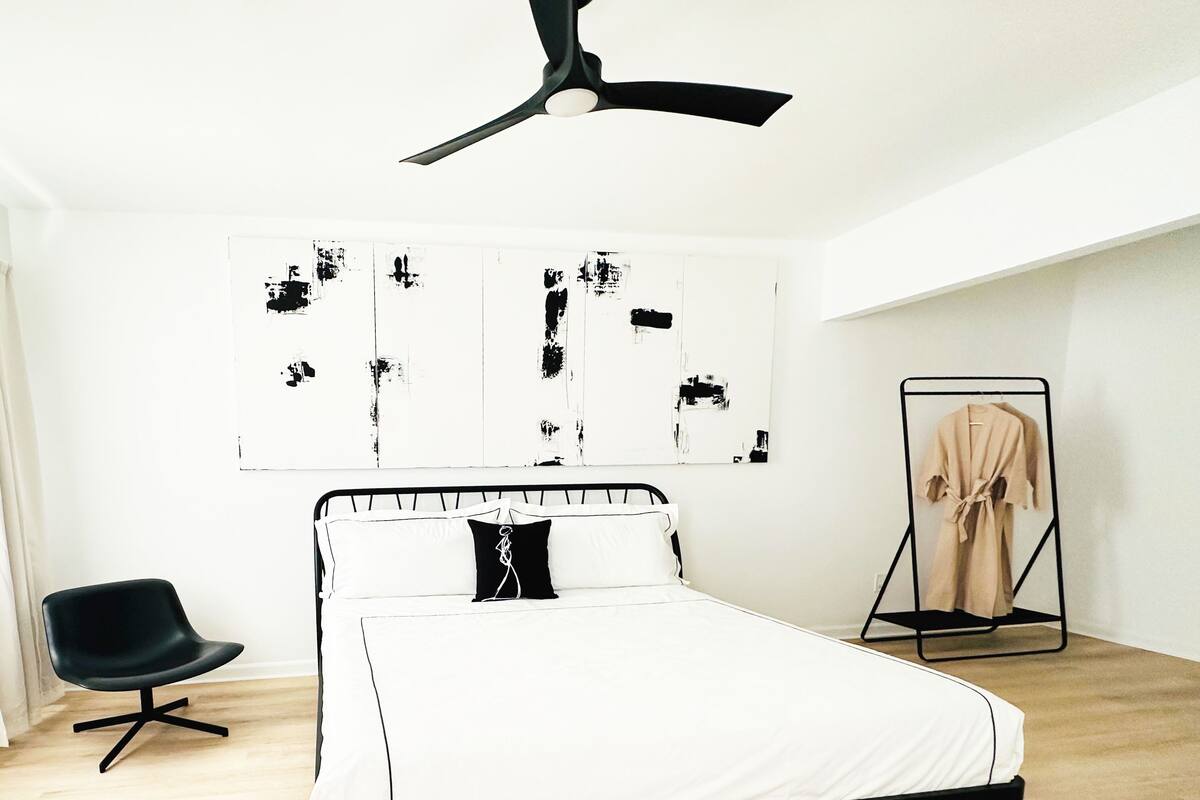 A bedroom with a bed, a chair, a ceiling fan, and abstract art on the wall.