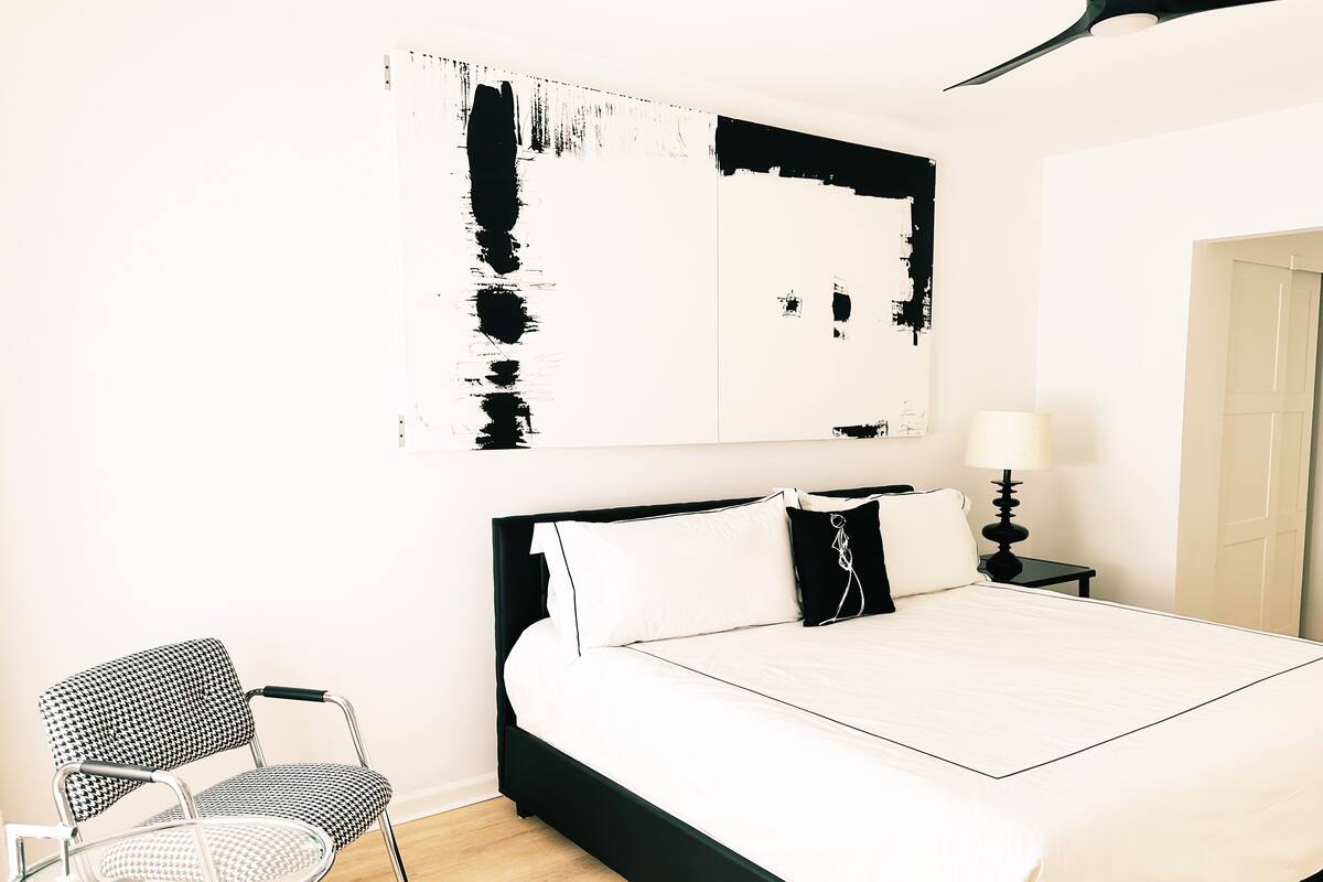 A modern bedroom with a large bed, a chair, a bedside table, and a wall art piece.