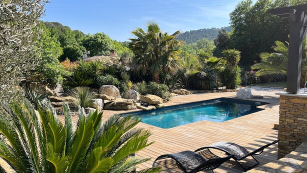 Un espace piscine doté d’une terrasse en bois, de chaises longues et d’une végétation luxuriante.