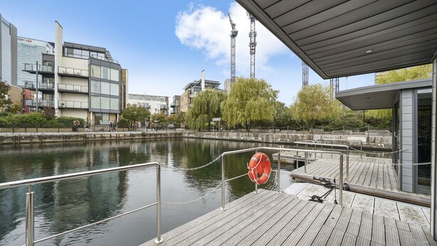 Un muelle de madera con un flotador salvavidas, un río tranquilo y edificios modernos al fondo.