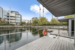 Une jetée en bois avec une bouée de sauvetage, une rivière paisible et des bâtiments modernes en arrière-plan.