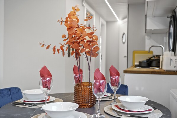 Une table à manger dressée avec de l’assiette blanche, des serviettes rouges et des verres transparents, et un vase décoratif placé au centre.