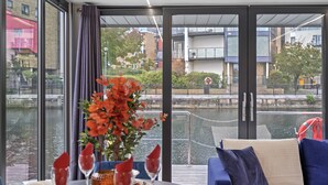 Een moderne eetruimte met een tafel gedekt voor twee personen, een bank en een groot raam met uitzicht op een rivier en gebouwen.