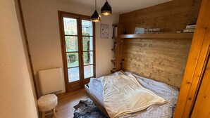 Un dormitorio con cabecera de madera, cama con ropa de cama estampada, un taburete blanco mullido, una alfombra y una ventana con vistas a la vegetación.