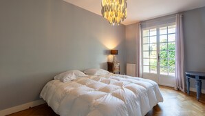 A bedroom with a large bed, a bedside table with a lamp, a window with curtains, and a chandelier.