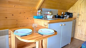 A cozy kitchen with wooden walls and a sloped ceiling. A wooden table with plates and a wine glass is set up. A sink and stove are visible in the background. - En Suite Glamping Pod, Sleeps 2 adults and 2 children, dog friendly. (Brodie)