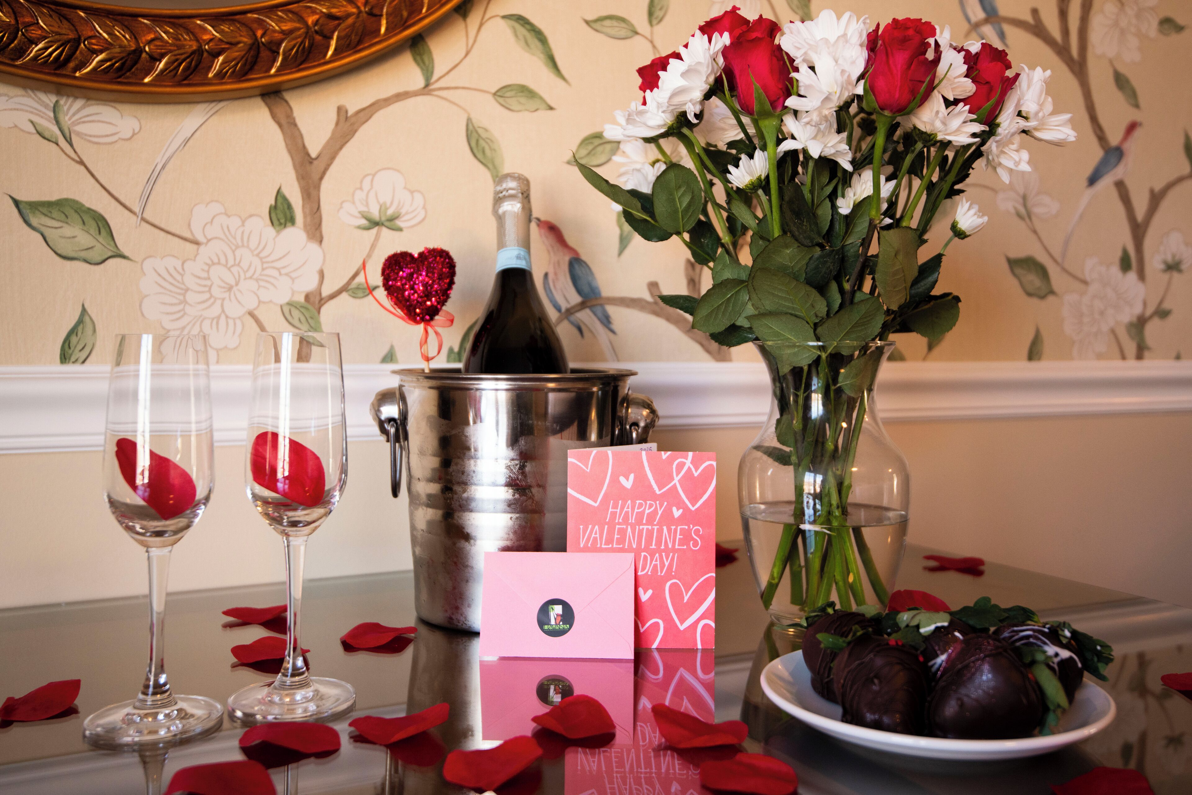 Taula amb una ampolla de xampany, un vas amb flors, un plat de xocolates i una targeta de Sant Valentí.