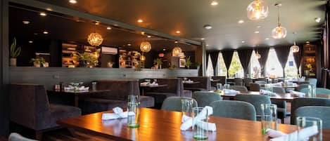 A restaurant with a modern interior, featuring wooden tables and chairs, hanging crystal chandeliers, and a view of the outdoors through large windows.