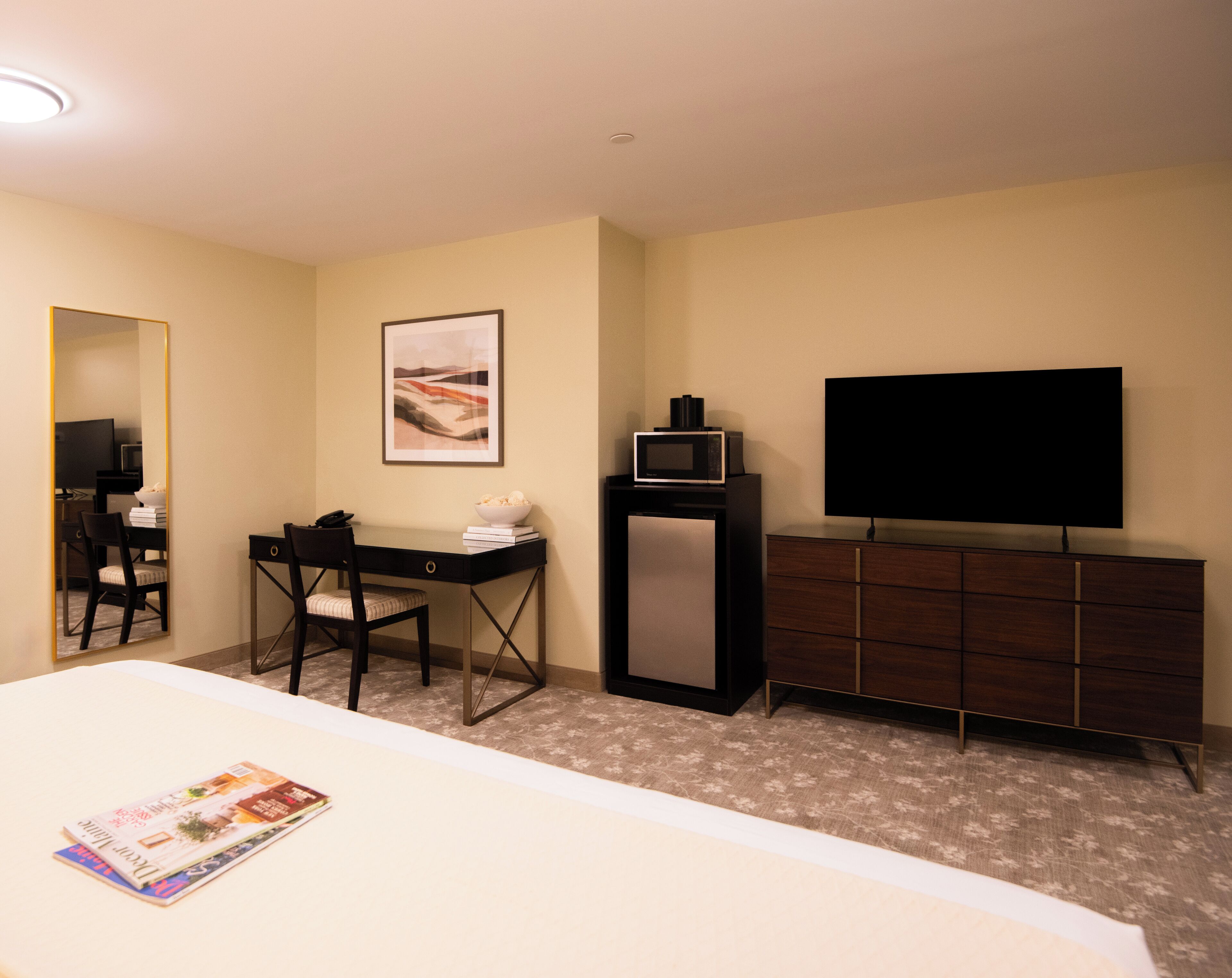 A hotel room with a bed, a desk with a chair, a television, a dresser, and a framed picture on the wall.