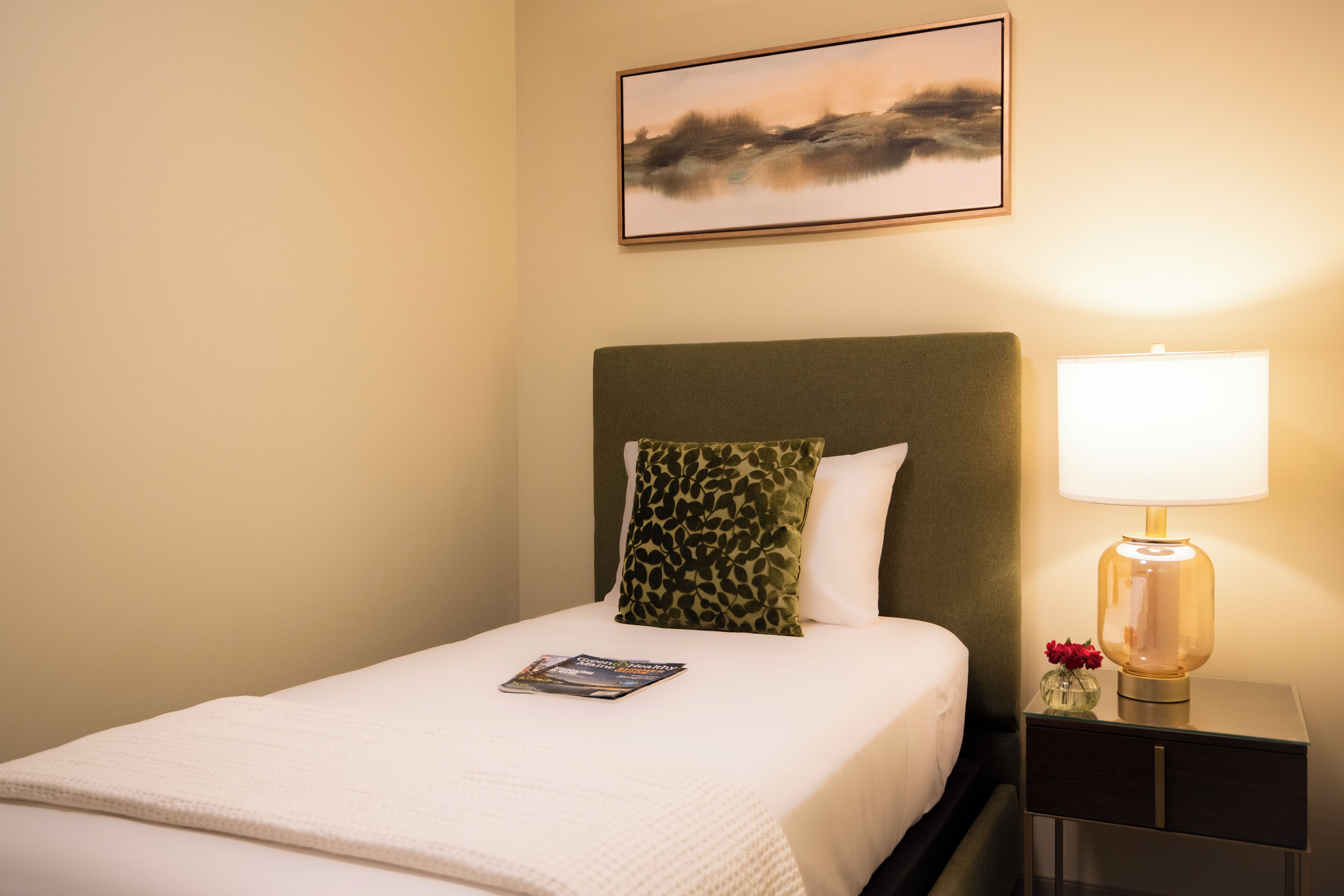 A neatly made bed with a magazine, a green patterned pillow, a lamp, and a framed picture on the wall.