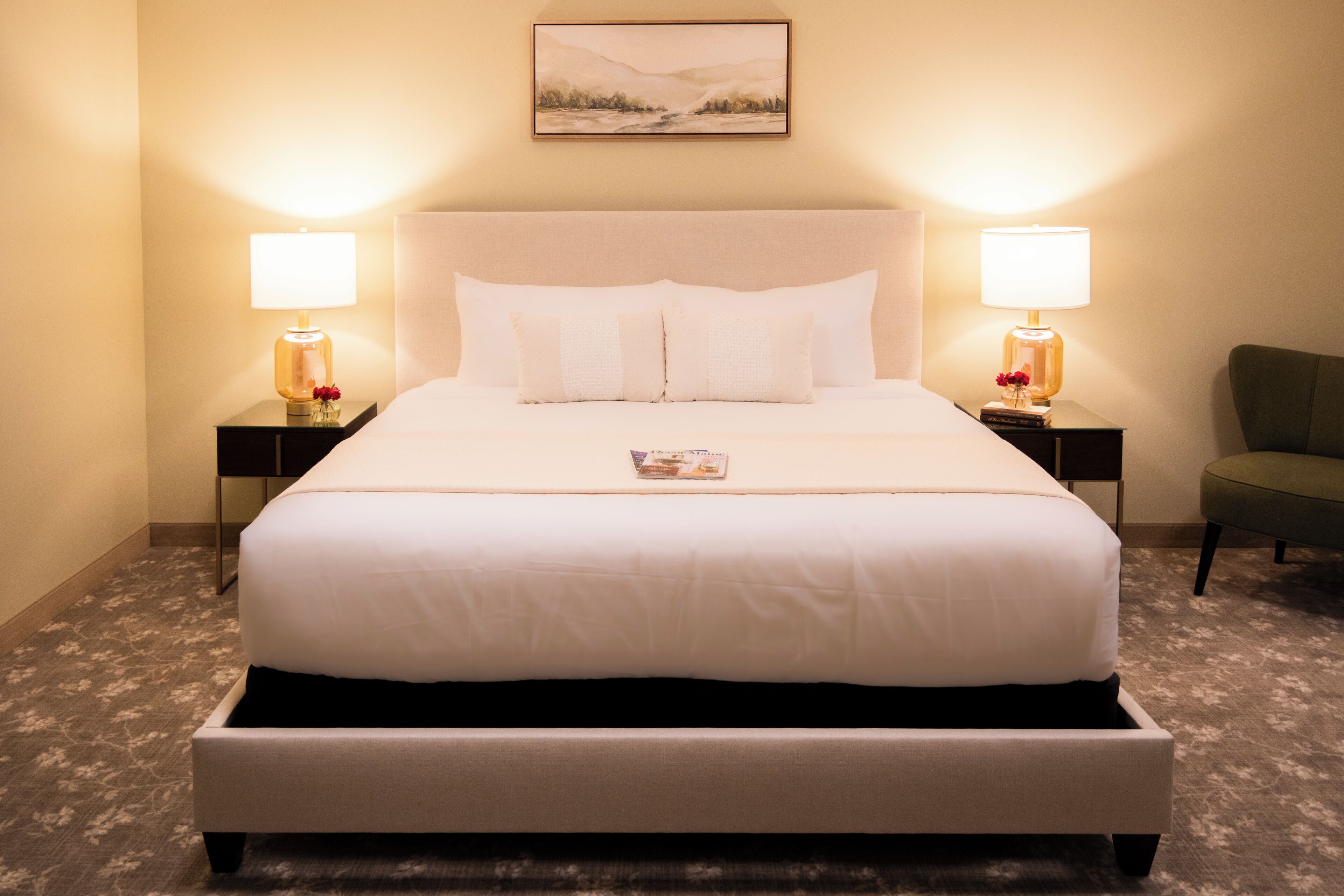 A neatly made bed with white linens, two bedside lamps, a framed picture on the wall, and a green chair in the corner.