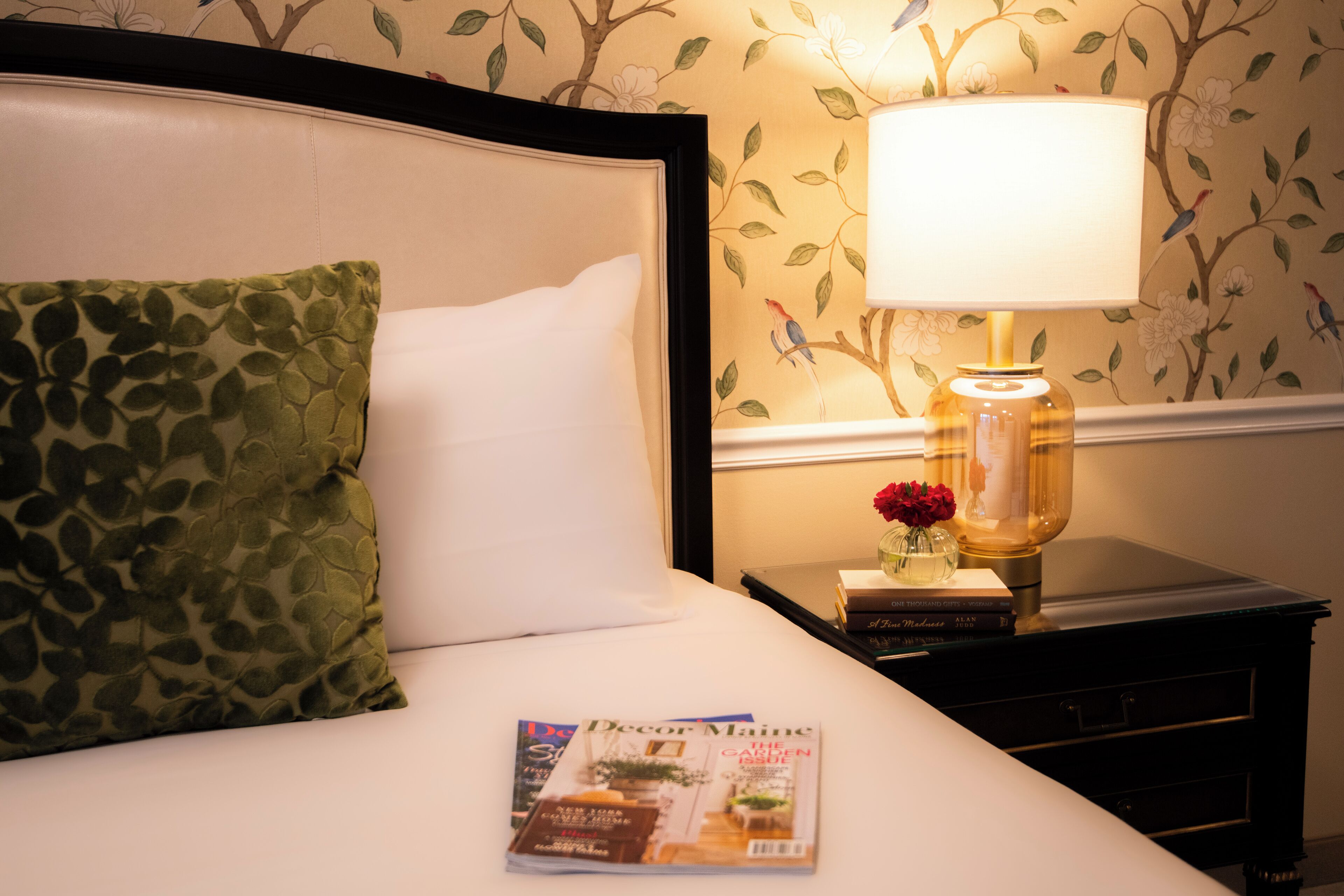 A neatly made bed with a green patterned pillow, a white pillow, and a magazine on the bed. There is a nightstand with a lamp, a vase with flowers, and a book.