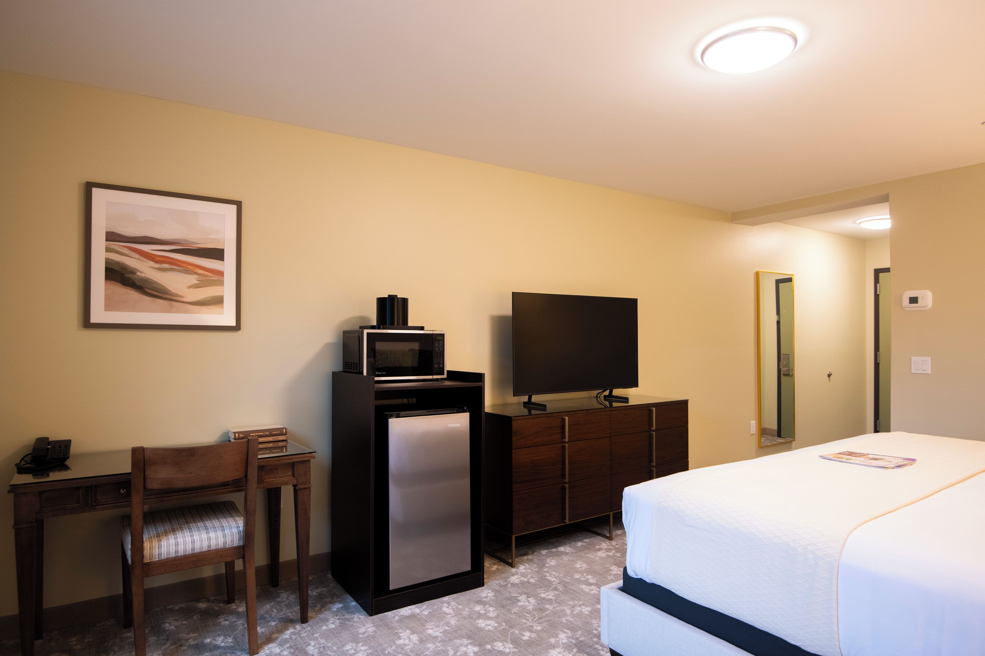 Una habitación de hotel con cama, escritorio con silla, refrigerador, microondas, televisión y un cuadro en la pared.