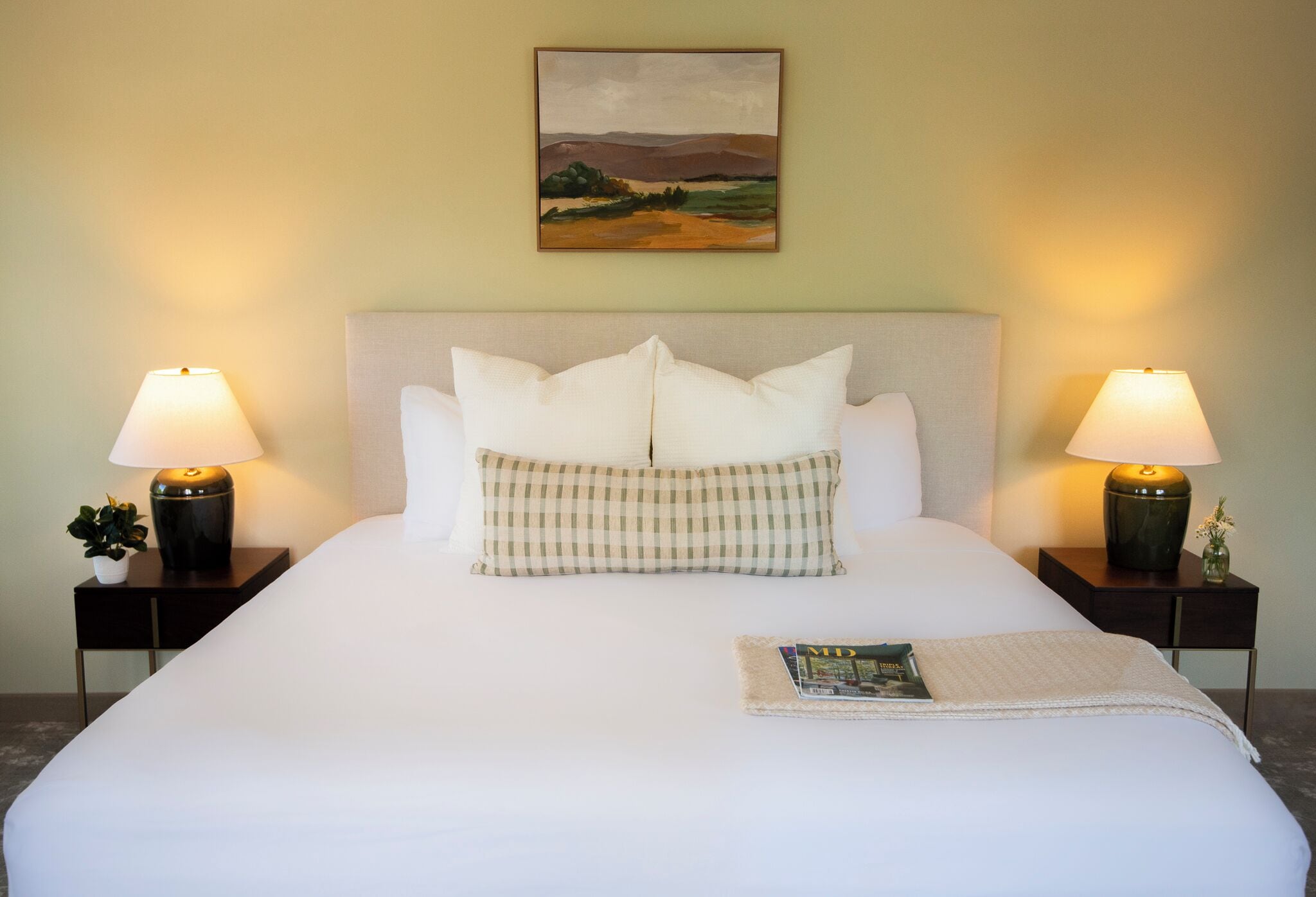 Un lit bien fait, avec du linge de lit blanc, deux lampes de chevet, un coussin à carreaux, un tableau représentant un paysage encadré et un magazine posé sur le lit.