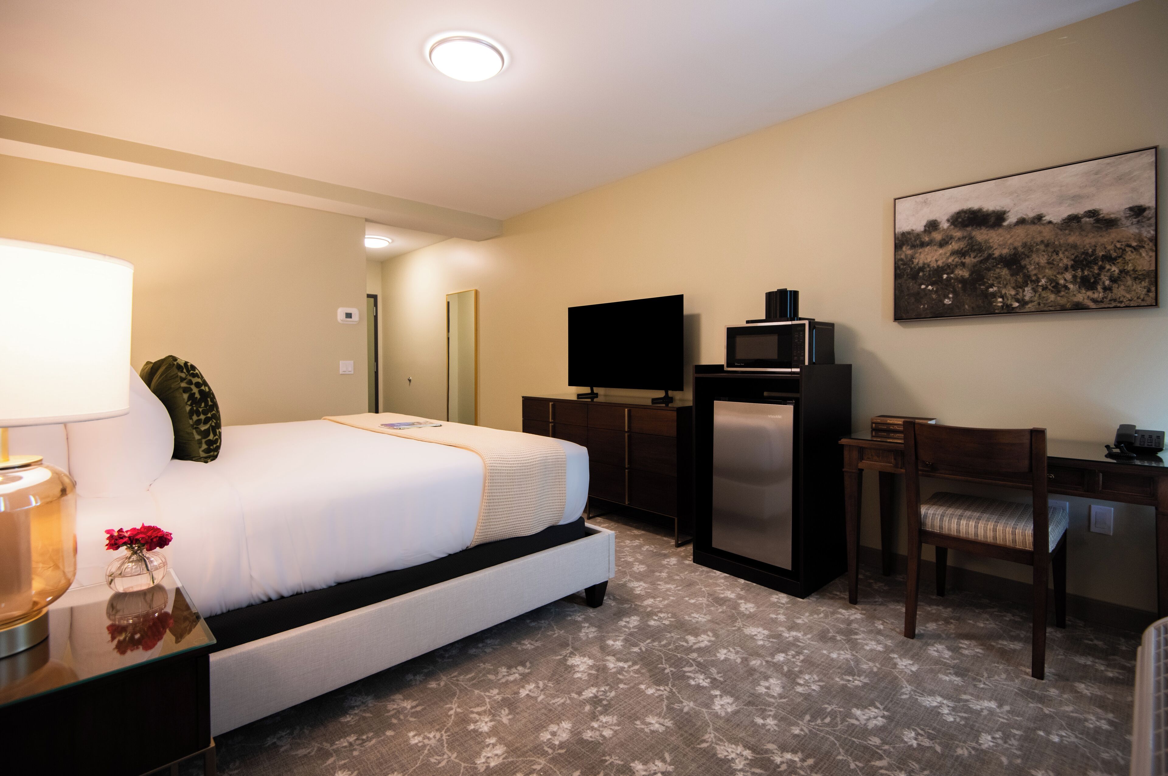 A hotel room with a bed, a desk, a television, a refrigerator, and a painting on the wall.