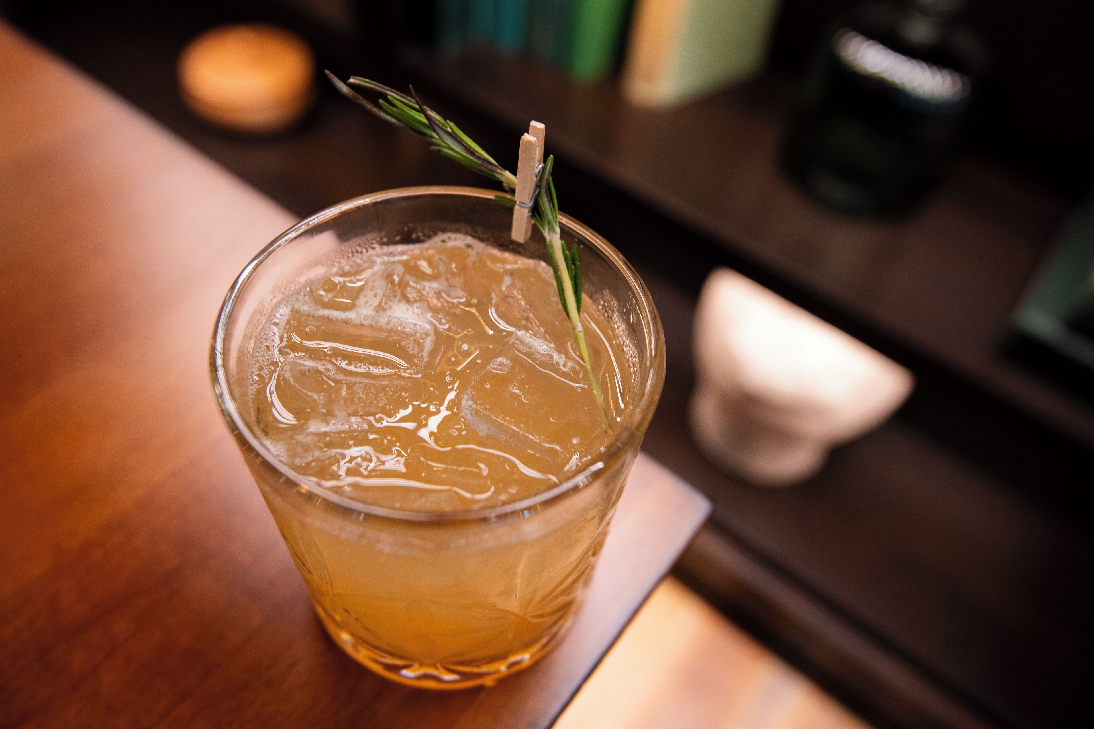 A glass of amber-colored beverage with ice cubes and a sprig of rosemary, placed on a wooden surface.