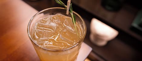 A glass of amber-colored beverage with ice cubes and a sprig of rosemary, placed on a wooden surface.