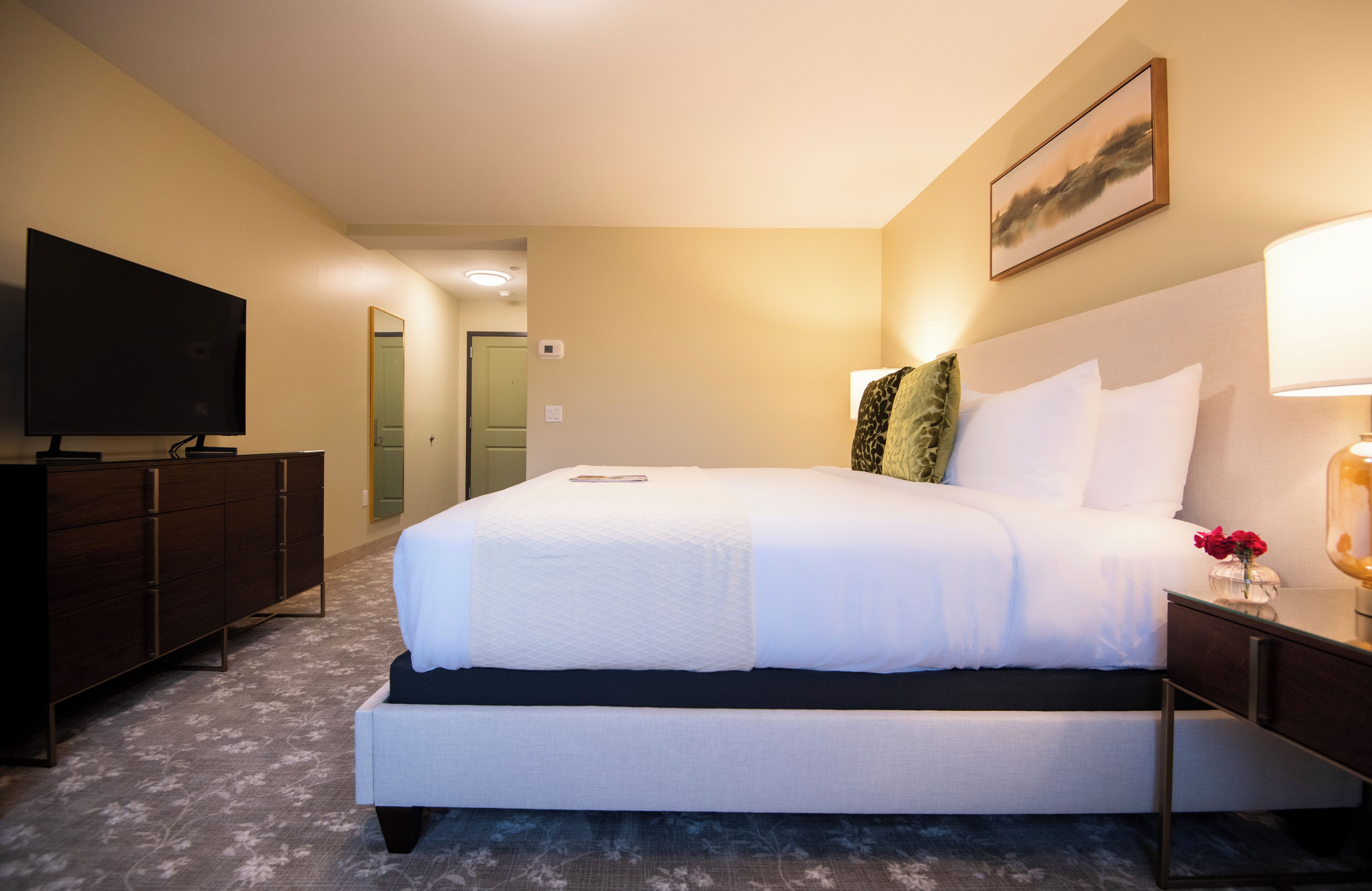 Ein Hotelzimmer mit einem großen Bett, einem Flachbildfernseher, einem Schminktisch und einem Nachttisch mit Lampe.