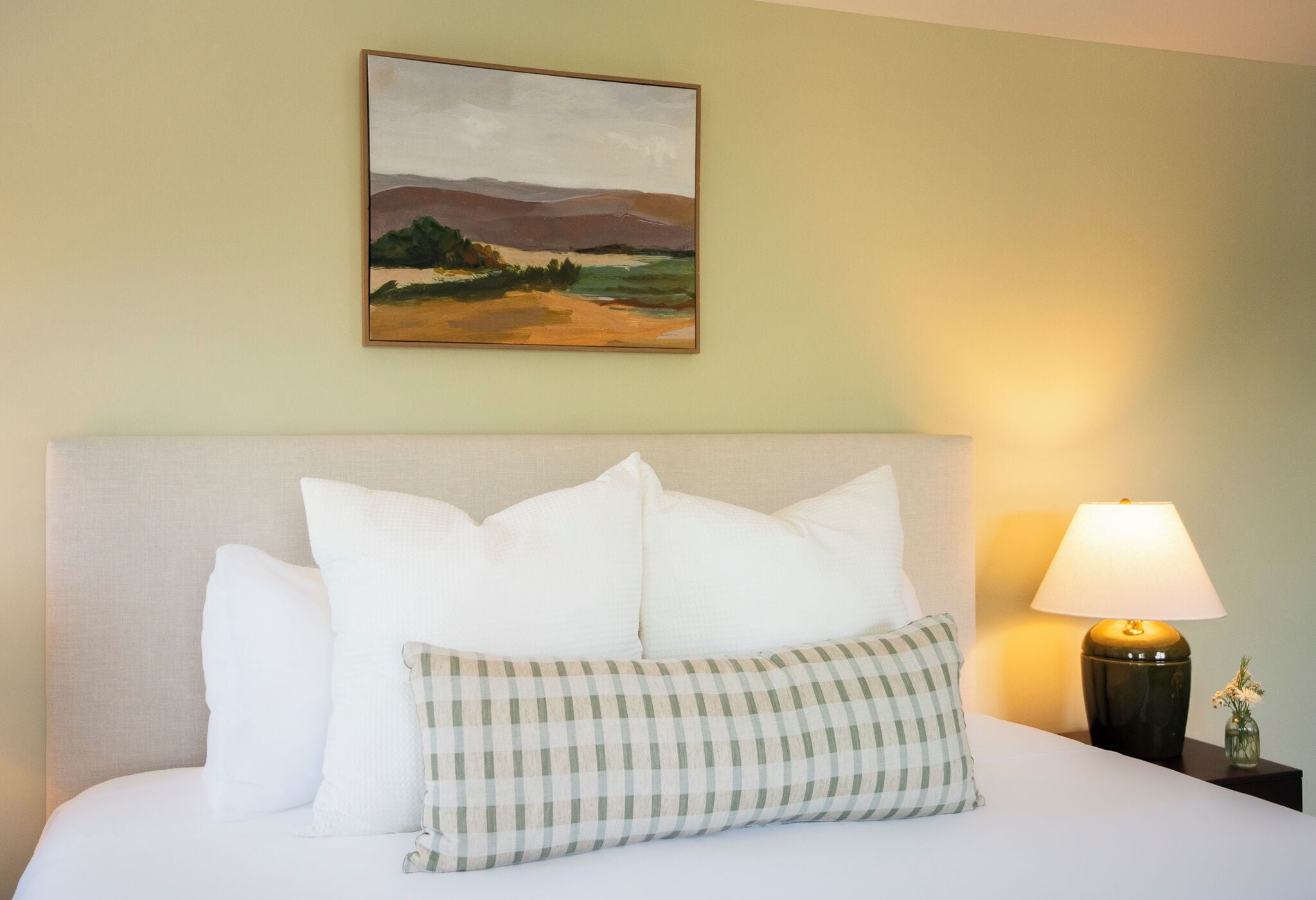 A neatly made bed with white and checkered pillows, a framed landscape painting above, and a bedside lamp on a nightstand.