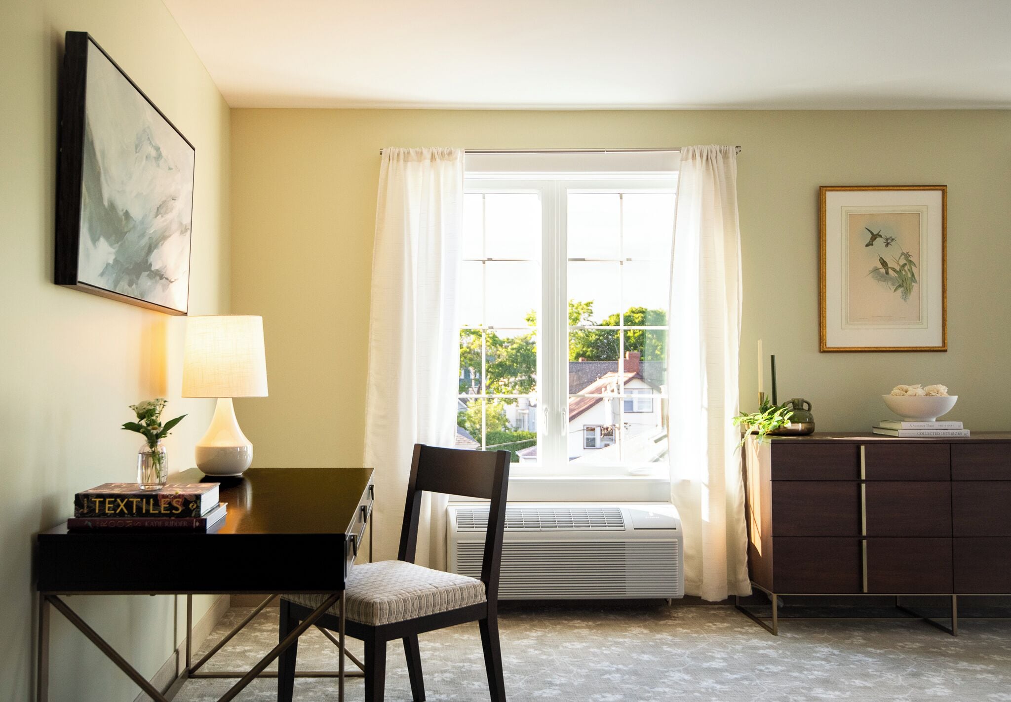 A room with a desk, chair, lamp, books, a radiator, and a window with curtains.