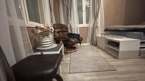 Une chambre à coucher avec un lit, une chaise, une petite table sur laquelle se trouve un vase, et une fenêtre avec des rideaux.