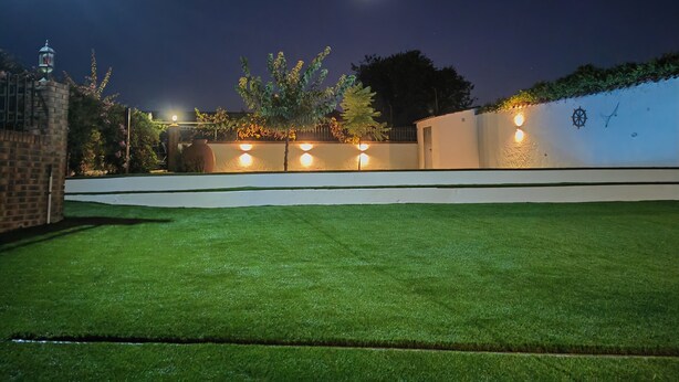 A well-lit outdoor area with a neatly trimmed lawn, a white wall with decorative lighting, and a brick structure on the left.