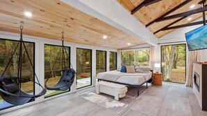A modern bedroom with a bed, a hanging chair, a bench, and a TV. - LUX Designer Private Waterfront Crooked River SEBAGO - GLASS WALLS - Huge Yard (Naples)