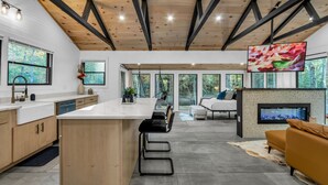 A modern kitchen with a central island, a dining area with a sofa and a TV, and a fireplace. - LUX Designer Private Waterfront Crooked River SEBAGO - GLASS WALLS - Huge Yard (Naples)