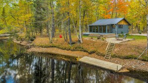 A cabin with a dock extending into a calm lake, surrounded by autumn-colored trees. - LUX Designer Private Waterfront Crooked River SEBAGO - GLASS WALLS - Huge Yard (Naples)