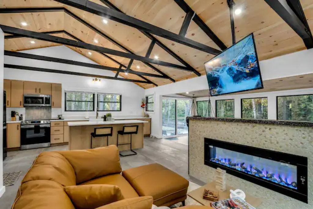 A modern kitchen with a central island, a dining area with bar stools, a living room with a sofa, and a fireplace.