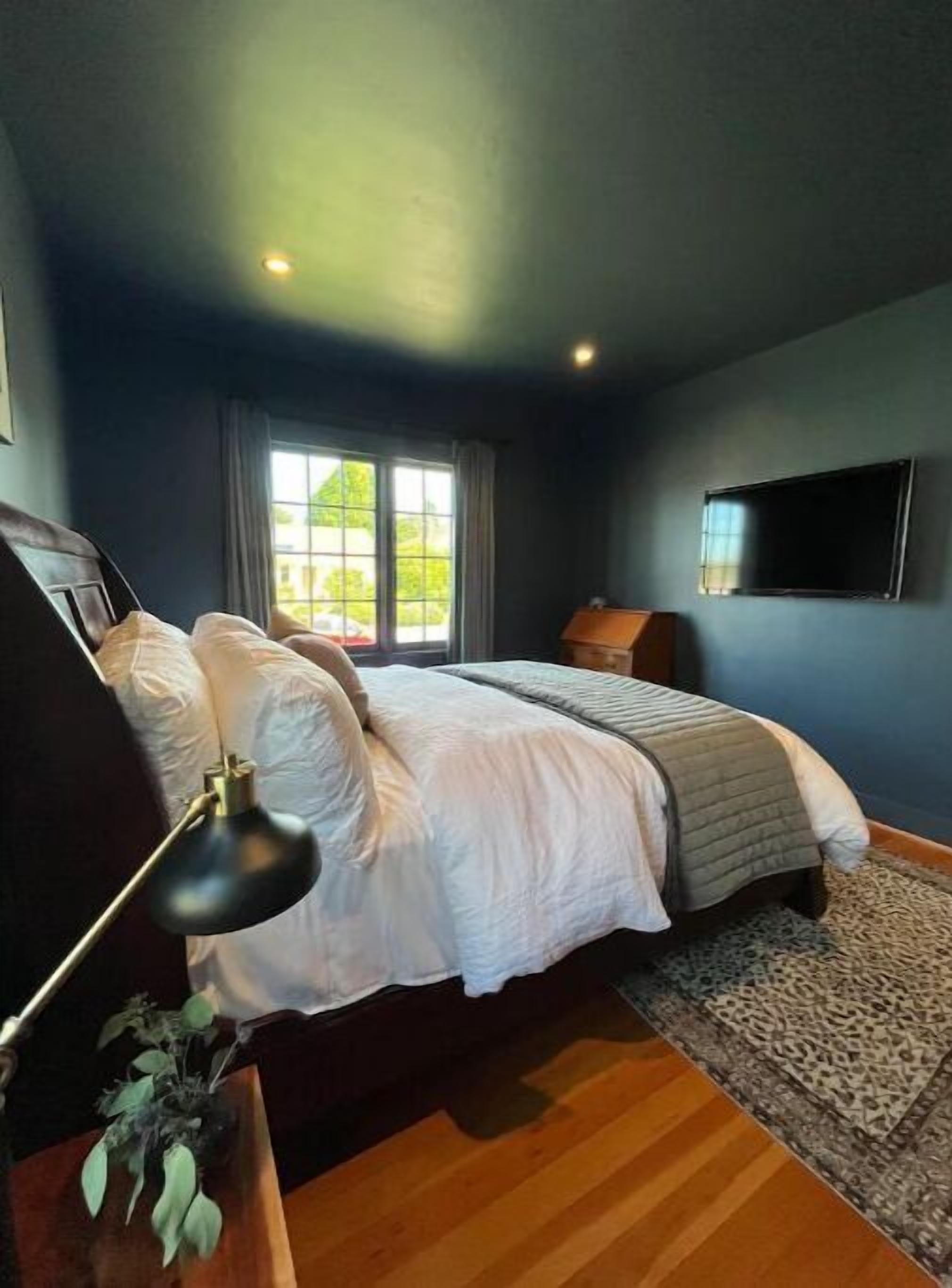 A bedroom with a bed, a desk with a lamp, a plant, and a window with curtains.