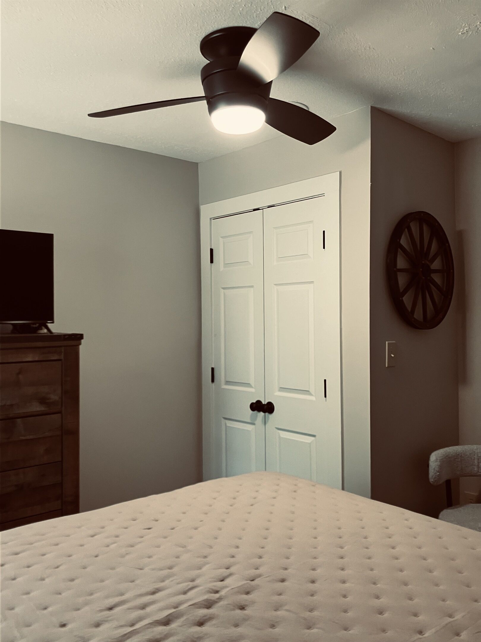 A bedroom with a bed, a dresser, a ceiling fan, a white door, and a wall-mounted round object.