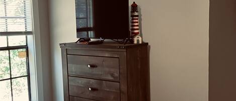 A bedroom with a bed, a dresser, a television on top of the dresser, and a window with blinds.