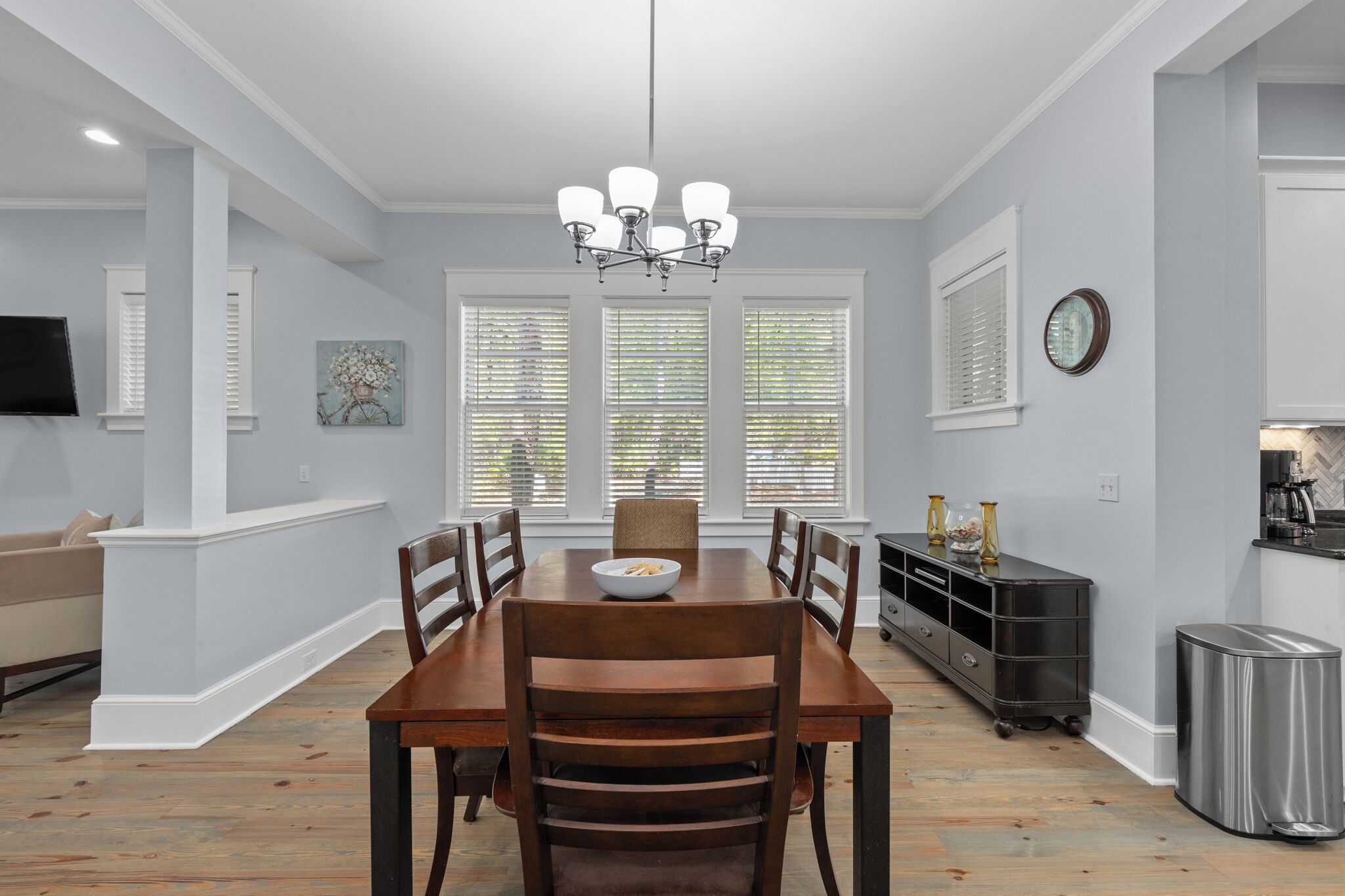 A dining room with a wooden table, chairs, a chandelier, and a kitchen area with a trash can.
